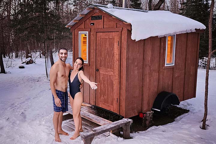 Young couple welcoming you into a cedar mobile sauna in a snowy Minnesota winter setting