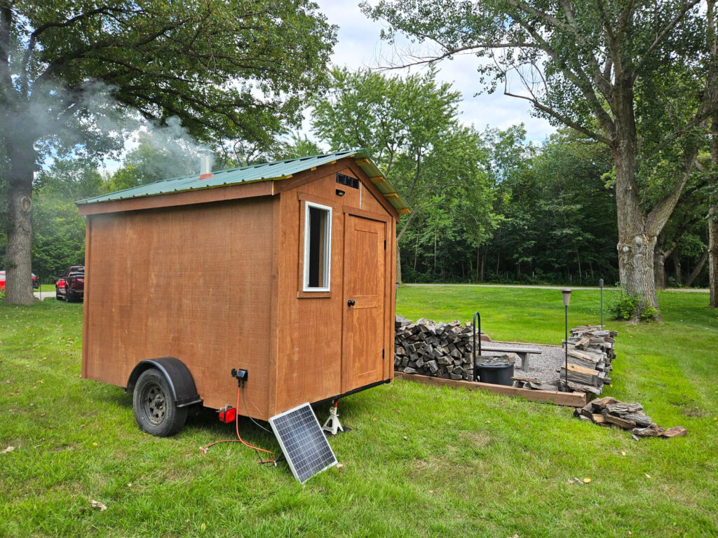 Cedar mobile sauna on a green lawn with steam rising, solar panel and woodpile visible